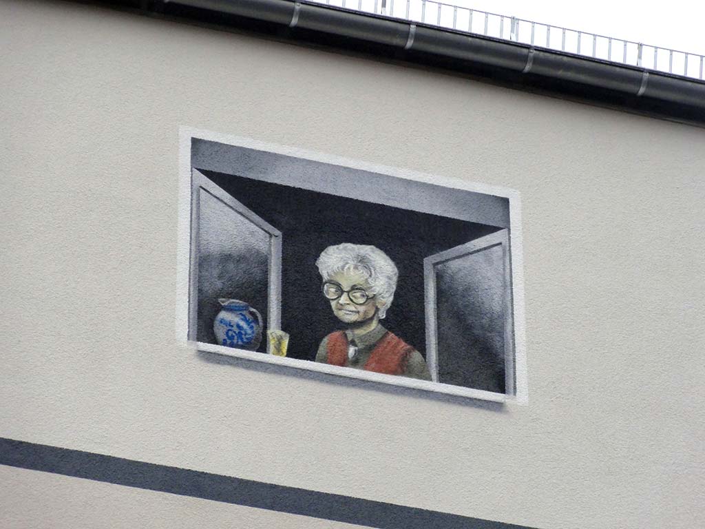 Geöffneter Reisverschluss gibt Blick frei auf Backsteinfassade, darüber schaut eine ältere Frau mit Bembel aus dem Fenster